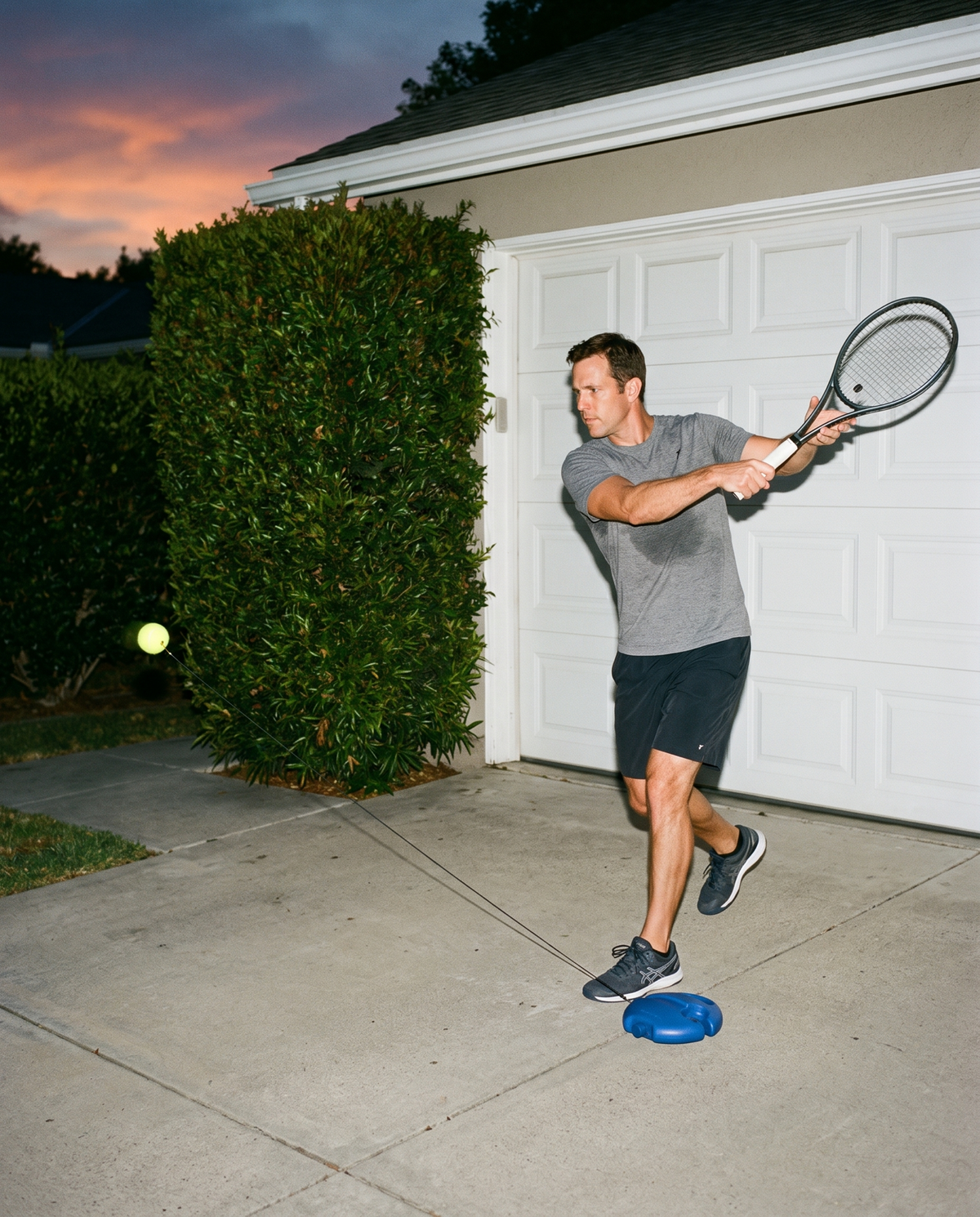 A man practicing forehands in his driveway at dusk with the RepCourt trainer base beside his feet